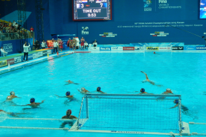 Eine Gruppe von Menschen spielt Wasserball in einem Pool mit einem Netz, umgeben von Zuschauern, während der FINA-Weltmeisterschaften 2015 in Kazan, mit Brettern, Trägern und einem Bildschirm mit Text und Logos.