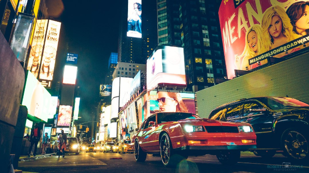 Roter Wagen fährt nachts auf einer belebten Stadtstraße in Times Square, New York City, mit beleuchteten Gebäuden, Werbetafeln und Verkehrszeichen.