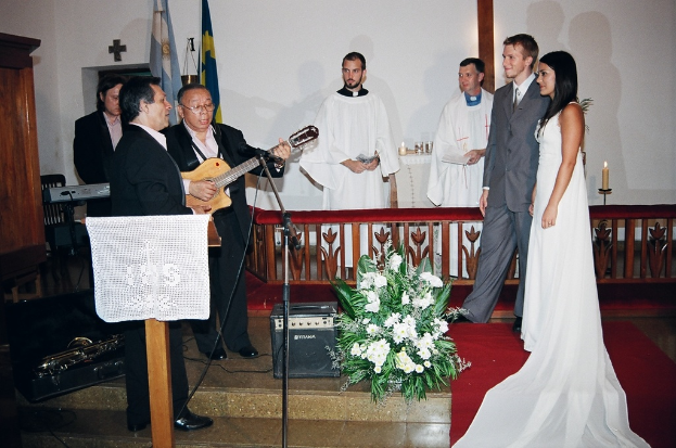 Eine HochzeitsSzene in einem Raum mit zwei Pfarrern, einer Braut, Bräutigam und drei Musikern; ein Podium mit Stoff vor den Musikern; ein Tisch mit Kerzen hinter den Pfarrern; eine Kerze in einem Halter hinter dem Paar; ein Bukett, Lautsprecher, Taschen und roter Teppich auf dem Boden; und Fahnen, eine Wand mit einem Kreuz, ein Schrank und ein Stuhl im Hintergrund.