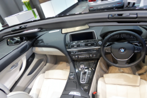 Das Innere eines BMW 6er Cabriolets mit Lederpolstern, einem Lenkrad, einem Armaturenbrett mit Knöpfen und Drehreglern, einem großen Touchscreen-Display, einem Seitenspiegel, einer Blume und zwei Autos im Hintergrund auf der Straße.