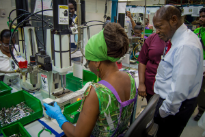 Eine Frau bedient eine Maschine mit einem Warnschild, umgeben von Drähten, Papieren und Gegenständen in einem grünen Behälter.