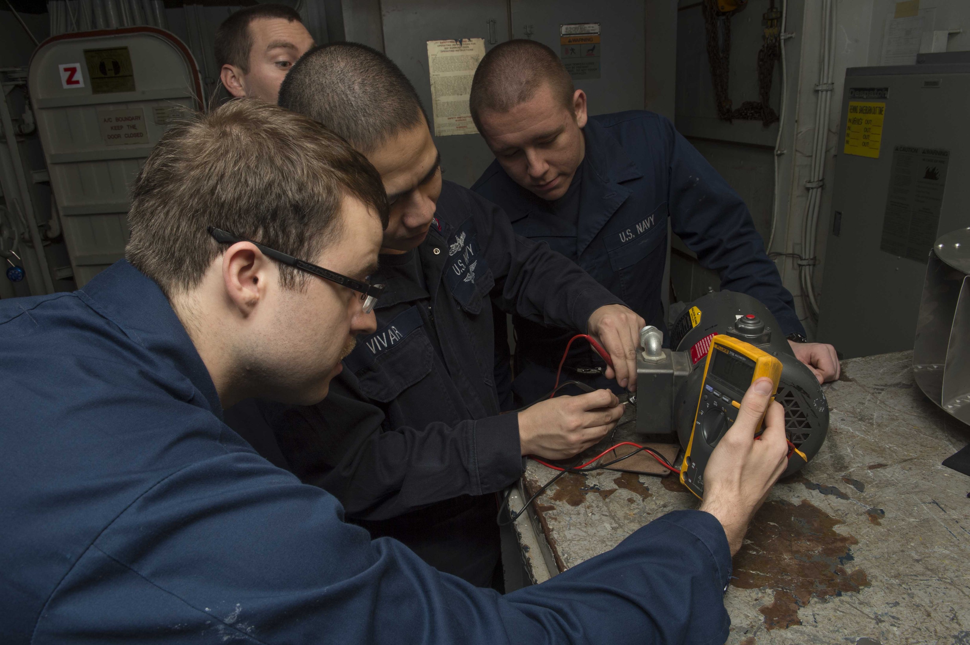 Vier Männer in einem Raum, die an einer Maschine arbeiten, mit einem Tisch voller Objekte rechts und Hintergrundmaschinen und Papieren an der Wand.