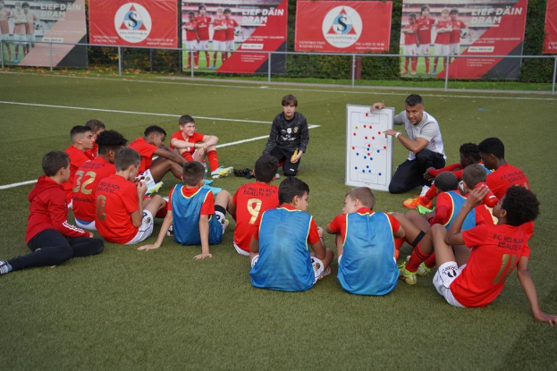 Junge Jungs auf einem Fußballfeld mit einer Tafel vor ihnen, einem Fußball in der Nähe und Bannern hinter ihnen an einem Zaun.