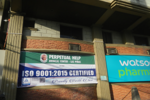 Ein Gebäude mit Säulen, Fenstern und Klimaanlagen, das ein Banner trägt, auf dem "Perpetual Help Medical Center - Las Pinas" und "ISO 9001 2015 zertifiziert" steht, sowie einen Pfahl und eine Kette vorne hat.