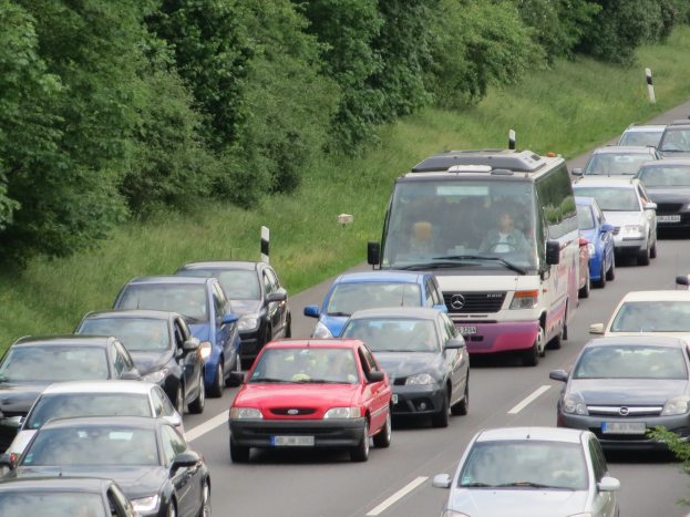 Ein Stau auf der Autobahn mit zahlreichen Autos und einem Van, mit sichtbaren Insassen, vor Bäumen und Gras im Hintergrund.