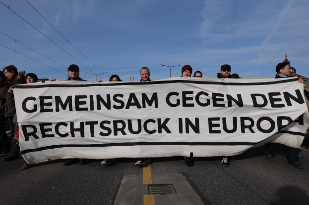 Eine Gruppe von Menschen auf der Straße mit einem Banner, auf dem "Gemeinsam Gegen den Rechtsruck in Europa" steht, im Hintergrund Straßenlaternen, Strommasten, Stromkabel, Gebäude und ein bewölkter Himmel.