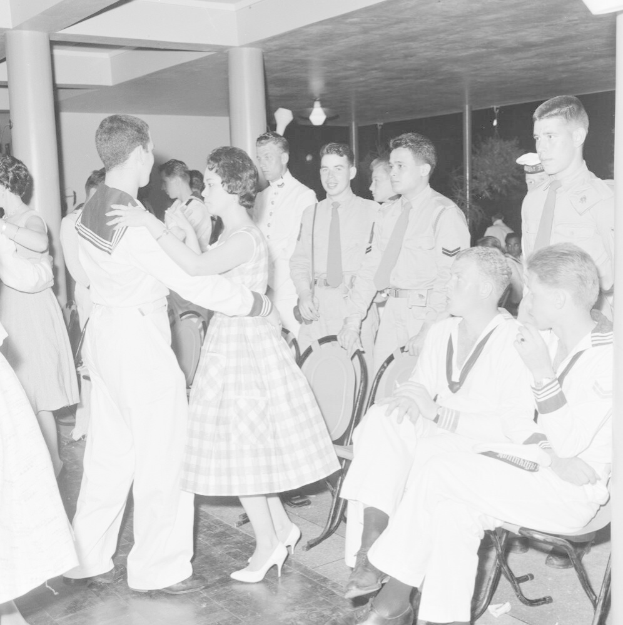 Schwarz-weißes Bild von Militärpersonal, das tanzt und sich vergnügt in einem dekorierten Saal mit Säulen und Beleuchtung.