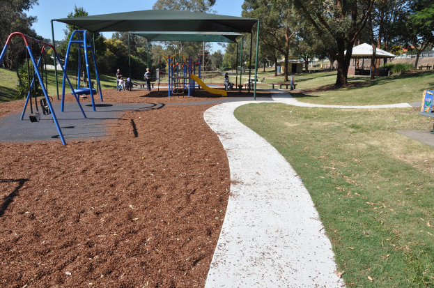 Ein Park mit einem Spielplatz mit Schaukeln, Rutschen und Bänken, umgeben von grünem Gras und Bäumen, mit Hütten und einem klaren blauen Himmel im Hintergrund.