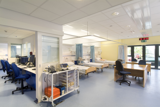 Krankenhauszimmer mit deckengehängten Lampen, Betten, Tischen, Stühlen, Monitoren, Tastaturen, Telefone, medizinischer Ausrüstung und Vorhängen an Glastüren.