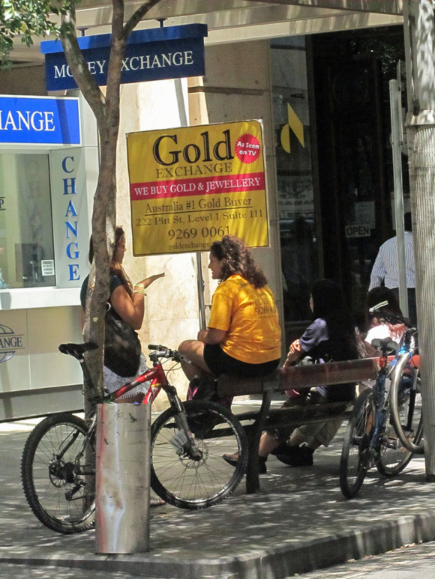 Eine Straßenszene mit Geschäften und Menschen in der Mitte, ein Fahrrad, eine Bank auf der rechten Seite und ein Baum auf der linken Seite.