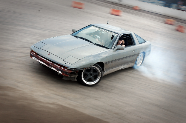Eine Person mit Helm fährt ein Auto auf einer Rennstrecke, wobei Rauch austritt, umgeben von orangefarbenen Objekten mit einem unscharfen Hintergrund, der hohe Geschwindigkeit andeutet.