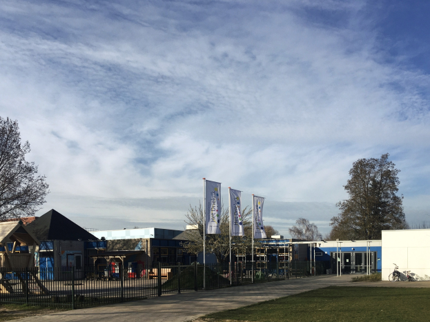 Großes Gebäude mit Metallzaun, Rasenfläche, Weg, Bäumen, Bannern, einem abgestellten Fahrrad, nahen Häusern und einem bewölkten Himmel, als neuer Spielplatz an einer Schule identifiziert.
