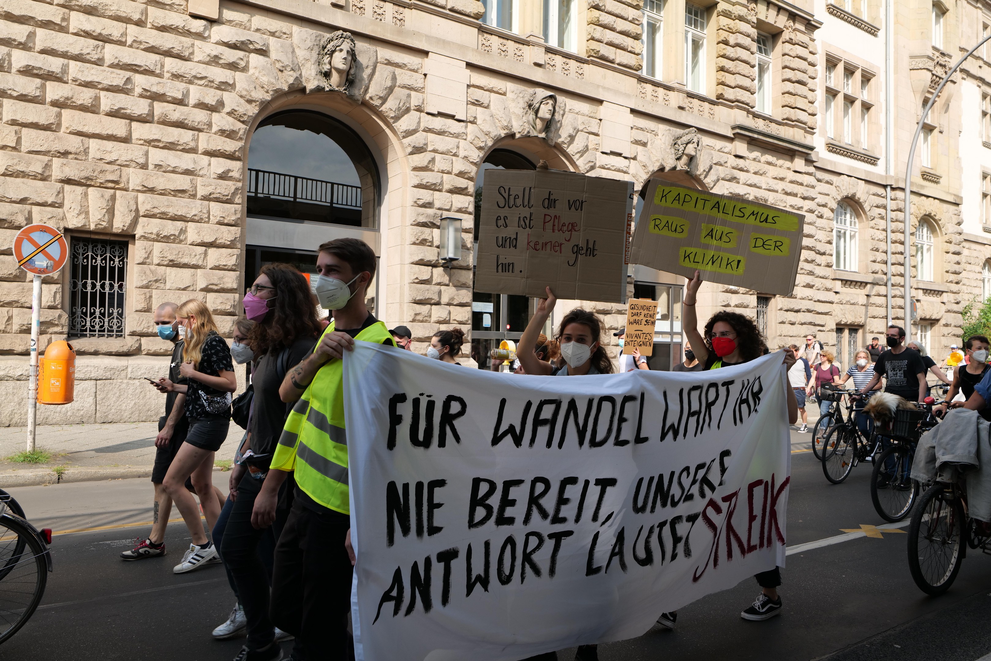 Eine Gruppe von Menschen marschiert bei einer Protestaktion gegen die Anti-Kriegsbewegung in Berlin, einige halten Schilder und andere fahren Fahrräder, im Hintergrund ist ein Gebäude mit Bögen, Säulen und Skulpturen zu sehen.