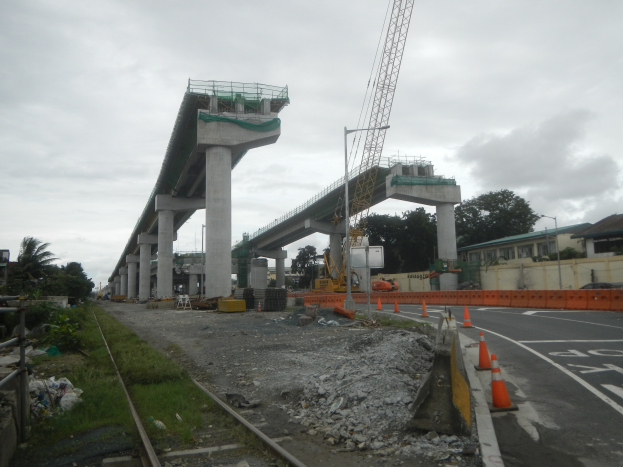 Baustelle mit einer Brücke im Hintergrund, eine Straße mit Verkehrskegeln auf der rechten Seite, Steine und Gras auf dem Boden, eine Bahnschiene auf der linken Seite, Bäume und Gebäude auf beiden Seiten der Straße und ein bewölkter Himmel.