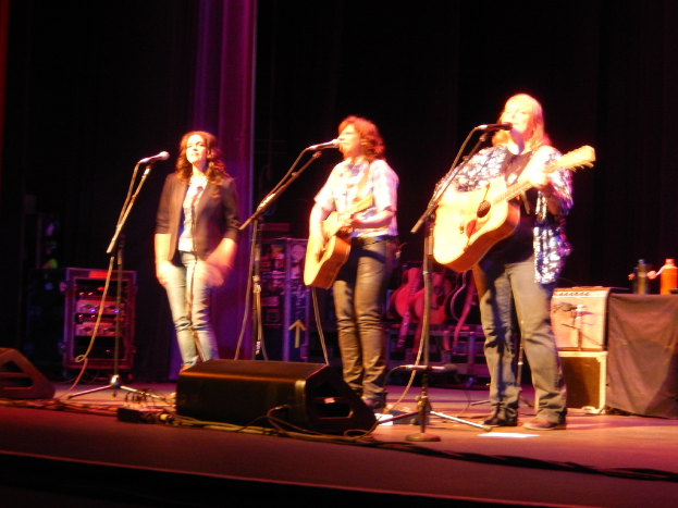 Drei Frauen performen auf der Bühne eines Musikkonzerts, jede singt in ein Mikrofon und spielt eine Gitarre, mit einem Tisch mit Wasserflaschen und zusätzlichen Gitarren dahinter.