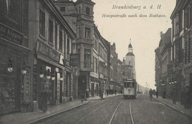 Schwarz-weißes Foto einer Straßenansicht in Brandenburg, Deutschland, mit einer Tram auf den Schienen, Passanten auf dem Gehweg, Gebäuden mit Fenstern, Schildern, Straßenlaternen und einem bewölkten Himmel.