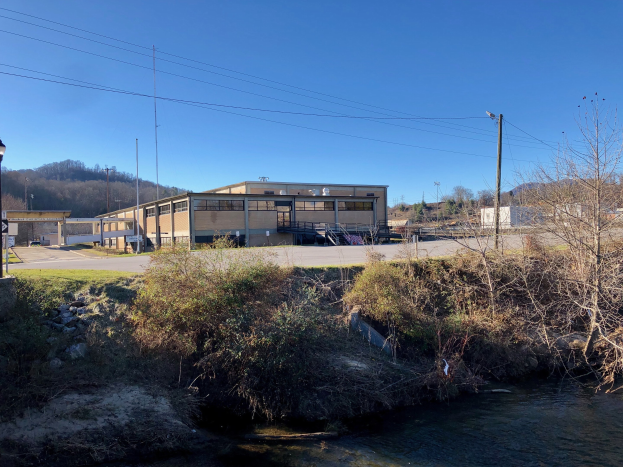 Ein Fluss fließt durch ein grünes Feld neben einem Gebäude, umgeben von Bäumen, Pflanzen, Gras, Steinen, Straßenlaternen, Strommasten, Stromkabeln, Hügeln und einem klaren blauen Himmel, mit dem Gebäude als Standort einer geplanten Wasseraufbereitungsanlage.
