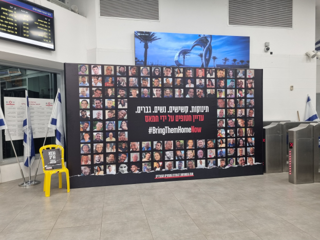 Eine große Wand zeigt zahlreiche Fotos, einen gelben Stuhl, israelische Regierungsflaggen auf Stangen, eine Tafel mit dem Text "Bring Them Home Now", einen montierten Bildschirm, Deckenleuchten und andere Gegenstände.