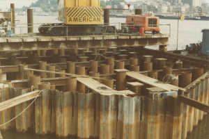 Baustelle mit einem Kran, der Stahlstäbe aus einem Haufen in der Nähe von Wasser hält, umgeben von Gebäuden, Bäumen und einem klaren blauen Himmel.