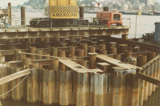 Baustelle mit einem Kran, der Stahlstäbe aus einem Haufen in der Nähe von Wasser hält, umgeben von Gebäuden, Bäumen und einem klaren blauen Himmel.