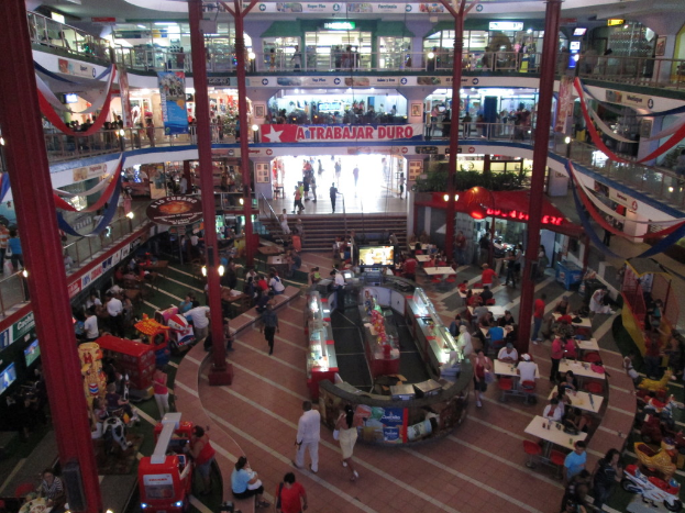 Ein belebtes Einkaufszentrum mit Säulen, Menschen, Tischen, Fliesen, Flaggen, Geschäften, einem Banner, Treppen und einem Namensschild.