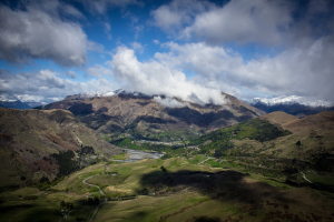 Ein atemberaubender Blick von der Spitze eines Berges in Queenstown, Neuseeland, mit saftig grünem Gras, Bäumen, einer gewundenen Straße und einem Himmel voller weißer, flauschiger Wolken.