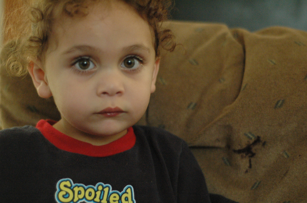 Ein Kind mit lockigen Haaren, das ein schwarzes Shirt mit der gelben Aufschrift "Spoiled" trägt und vor einem braunen Stoffhintergrund steht.