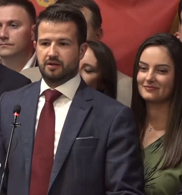 Ein Mann im Anzug und mit Krawatte steht an einem Rednerpult mit einem Mikrofon und spricht vor einem Publikum mit einem Banner im Hintergrund, wahrscheinlich in Reaktion auf ein Regierungshandelsverbot.