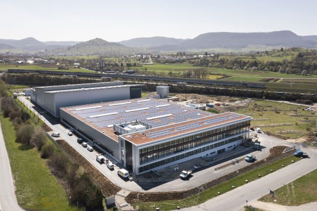 Luftaufnahme eines großen Industriegebäudes mit Solarmodulen auf dem Dach, umgeben von Fahrzeugen, Bäumen, Gras und einem klaren blauen Himmel im Hintergrund.