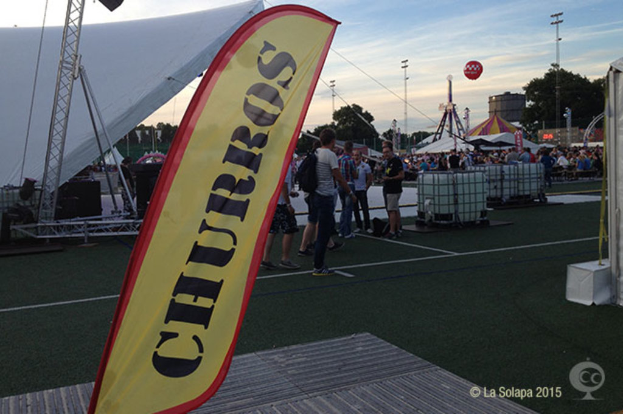 Eine Karnevalsszene mit Zelten, einem zentralen Mast, Menschen, Bäumen, Strommästen, Straßenbahnen und einem eisernen Ständchen auf der linken Seite unter einem Himmel.