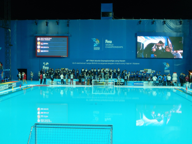 Gruppe von Menschen, die um ein Hallenbad mit Netz am Boden stehen, mit einer Wand im Hintergrund, die "FINA World Championships 2015 Kazan"-Text und Logos anzeigt, sowie Tr├Ąger und Lichter