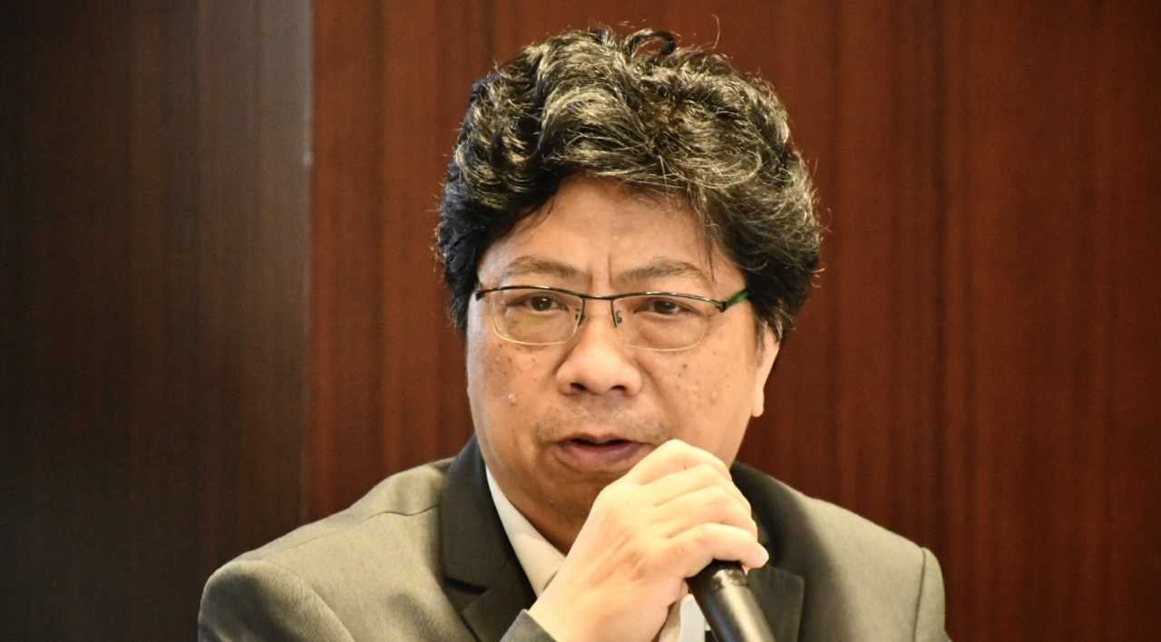 Ein Mann im Anzug mit einem Mikrofon, der lebhaft vor einer Holzwand spricht.