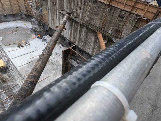 Eine Baustelle mit einem großen Metallrohr in der Mitte, umgeben von Arbeitern, hölzernen Gegenständen und diverser Ausrüstung, mit Fahrzeugen und einem Gebäude im Hintergrund.
