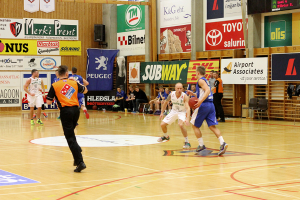 Ein Basketballfeld mit Spielern im Vordergrund und einer hölzernen Wand mit Postern und Werbetafeln im Hintergrund.