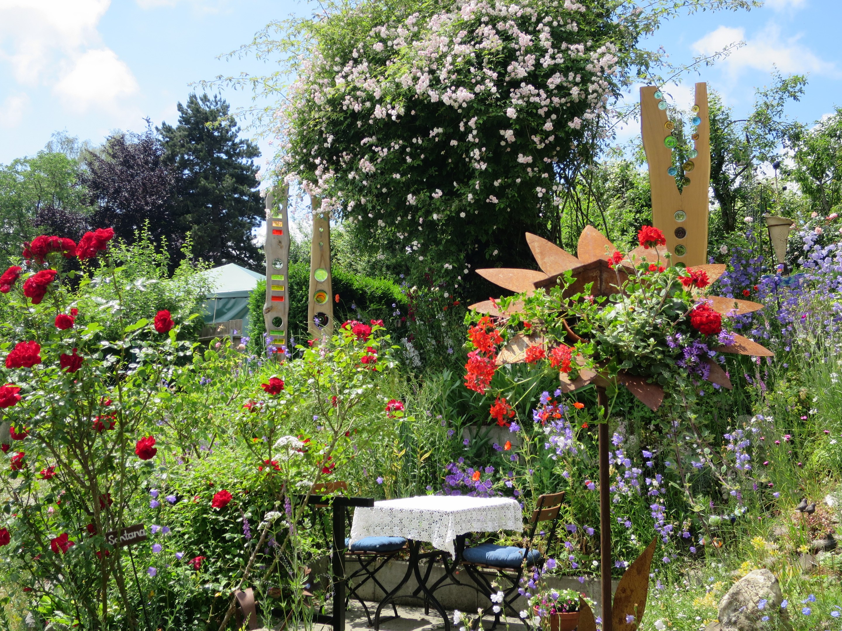 Ein Garten auf der Chelsea Flower Show mit bunten Blumen und Pflanzen, einem Tisch mit einer Decke und Stühlen in der Mitte, umgeben von Bäumen unter einem bewölkten Himmel.