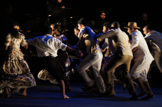 Eine Gruppe von Menschen tanzt im Vordergrund, während im Hintergrund eine weitere Gruppe von Menschen, Stände und Mikrofone zu sehen sind, alles auf einem Boden.