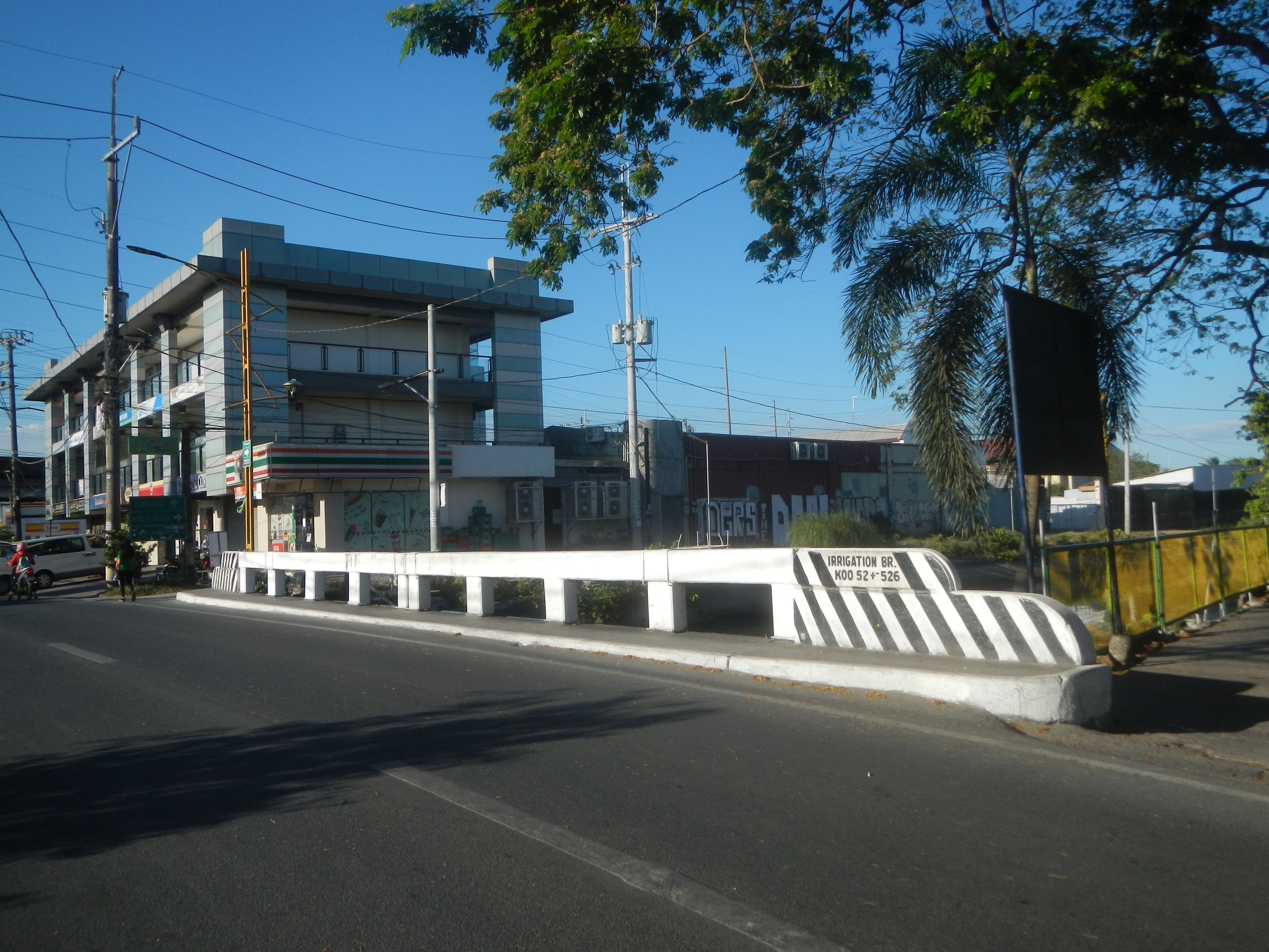 Stadtstraße mit Gebäuden, Strommasten, Kabeln, Fahrzeugen, Absperrpollern, Namensschildern, Bäumen, Pflanzen und einem klaren blauen Himmel.