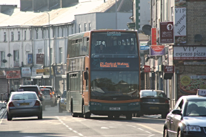 Eine Straße mit Autos und einem Bus, Gebäude mit Wänden, Fenstern, Tellern und Dächern, Plakate und Banner an den Wänden und ein Pfahl mit einer Straßenlaterne.