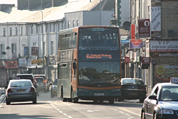 Eine Straße mit Autos und einem Bus, Gebäude mit Wänden, Fenstern, Tellern und Dächern, Plakate und Banner an den Wänden und ein Pfahl mit einer Straßenlaterne.