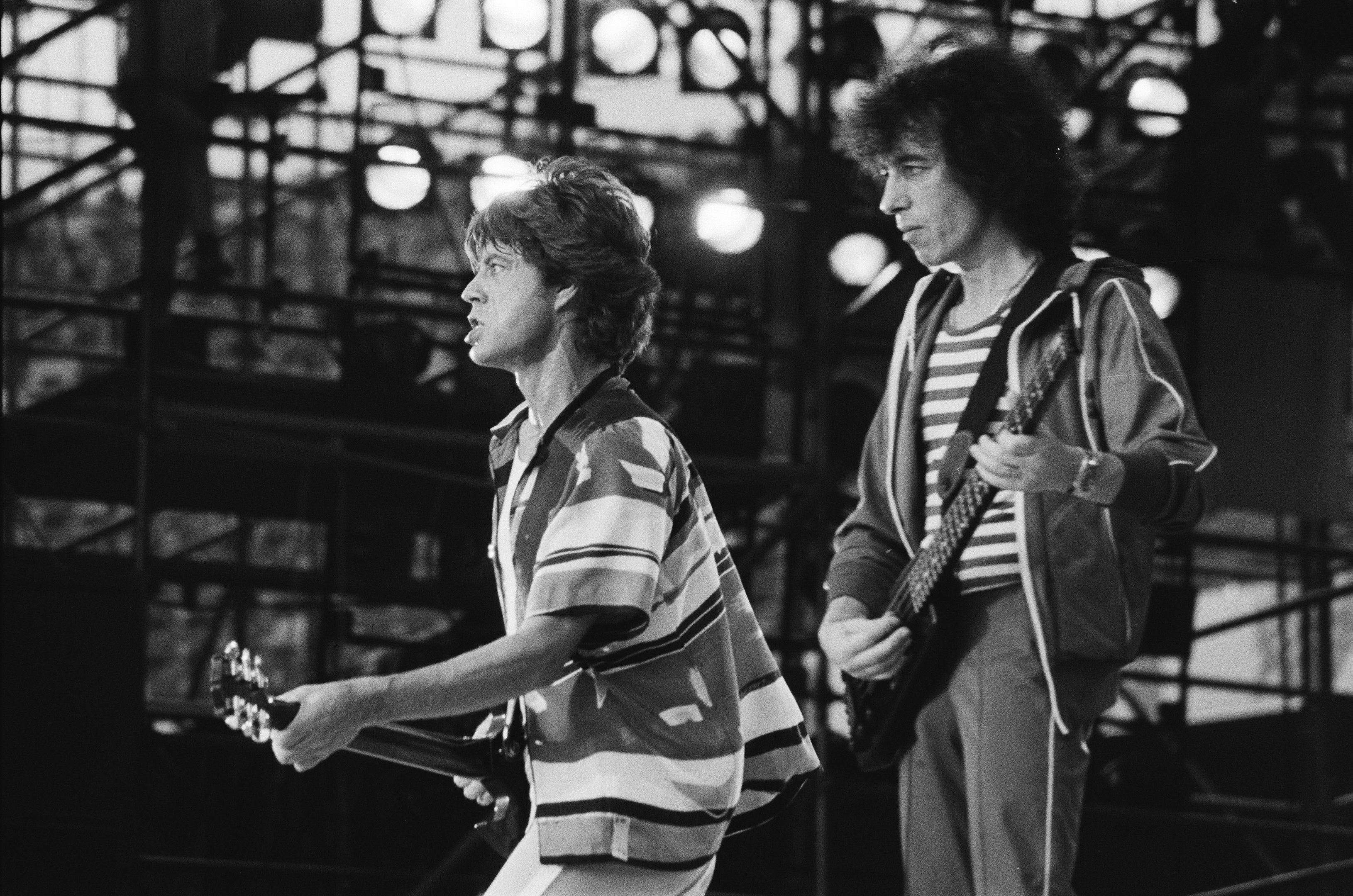 Schwarzes und weißes Foto von Mick Jagger und Brian May bei einem Auftritt auf der Bühne im Hammersmith Odeon, beide spielen Gitarren unter hellen Bühnenlichtern.