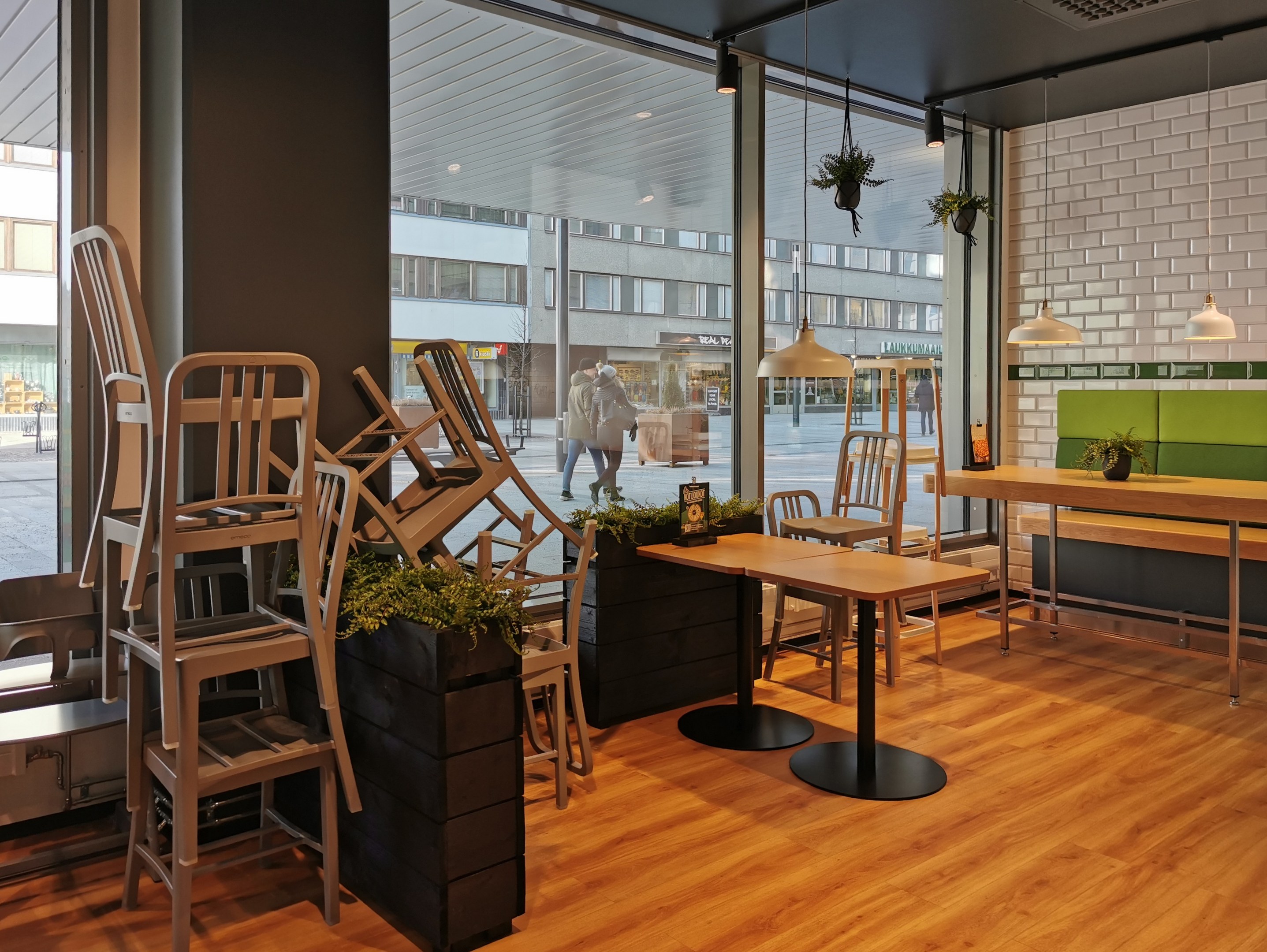 Ein Berliner Restaurant mit Holztischen und -stühlen in der Nähe eines großen Fensters, das eine städtische Außenansicht mit Fußgängern, Gebäuden und Straßenelementen zeigt.