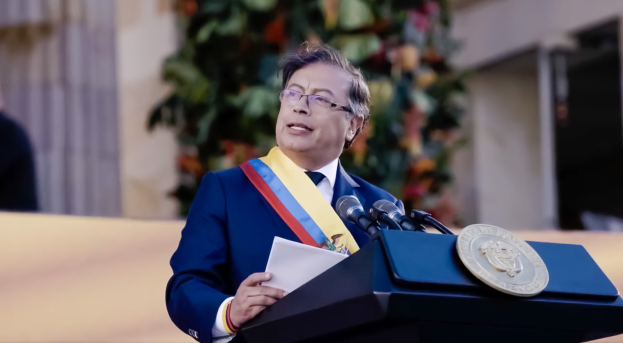 Ein Mann in einem blauen Anzug und Krawatte steht an einem Rednerpult, hält Papiere und spricht in ein Mikrofon, mit einer Pflanze und einer Wand dahinter, was auf eine formelle Umgebung hinweist.