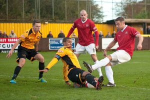 Fußballspieler in blauen und roten Trikots spielen auf einem Rasenfeld mit einem Ball, während Zuschauer außerhalb des Spielfelds stehen und sie anfeuern, mit einem Baum und Himmel im Hintergrund.