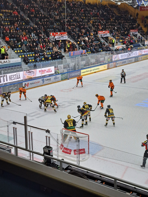 Eine Gruppe von Menschen, die Hockey auf einer Indoor-Eisfläche mit einem Netz und einer Umzäunung spielt, beobachtet von Zuschauern auf Stühlen, mit Bannern und Überdachungsbeleuchtung im Hintergrund.
