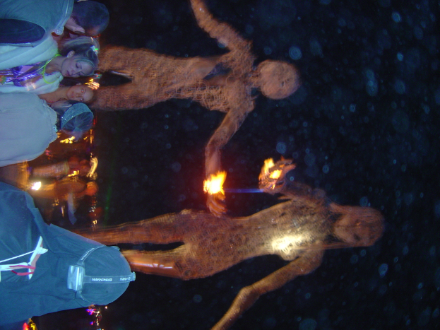 Menschen auf der linken Seite des Bildes mit brennenden Statuen dahinter und Lichter im Hintergrund.