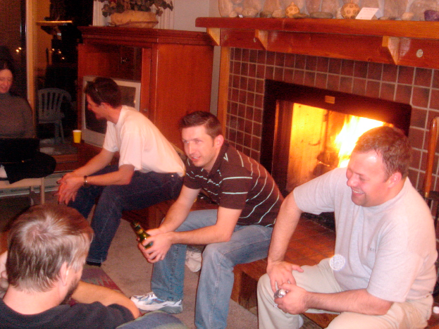 Vier Männer sitzen auf einer Bank, eine Frau sitzt links auf einem Stuhl, ein Feuer brennt im Kamin, ein Holzbrett hält Papier, einen Ball und Showstücke, ein Holztisch hält einen Blumentopf und eine Tasse, mit einem Fernseher, Stuhl und Lampen im Hintergrund.