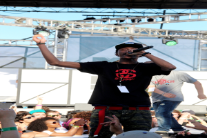 Eine Person in einem schwarzen T-Shirt hält ein Mikrofon auf einer Bühne mit Menschen drumherum, während eine andere Person in einem grauen T-Shirt im Hintergrund unter einem blauen Himmel steht.