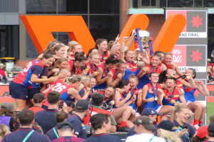 Eine Gruppe von Frauenfußballspielerinnen in Sportuniformen feiert mit einem Pokal vor einem Publikum, mit einem Gebäude mit Glasfenstern und einer Texttafel im Hintergrund.