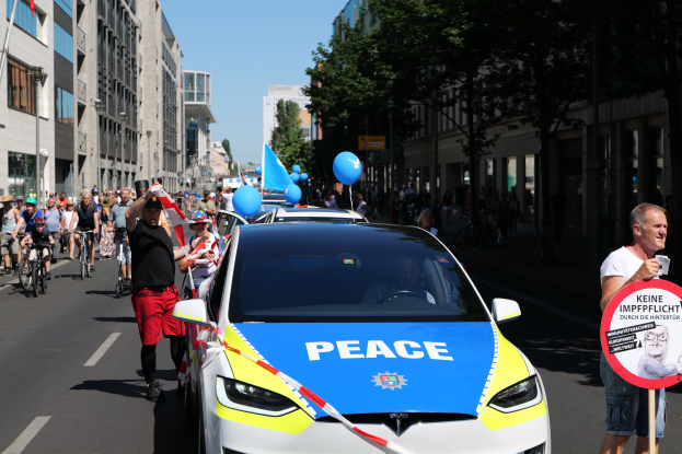 Eine Gruppe von Menschen, die auf Fahrrädern eine Straße entlangfahren, mit einem Polizeiauto im Vordergrund, das Text anzeigt, und einer Person, die eine Tafel mit einem Bild und Text hält, vor einem Hintergrund aus Bäumen, Gebäuden mit Fenstern, Laternenpfählen und einem klaren blauen Himmel.