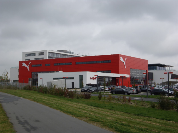 Außenansicht der Puma-Fabrik in Deutschland mit Gebäuden, Fahrzeugen, Straßeninfrastruktur, Grünflächen und einem bewölkten Himmel.